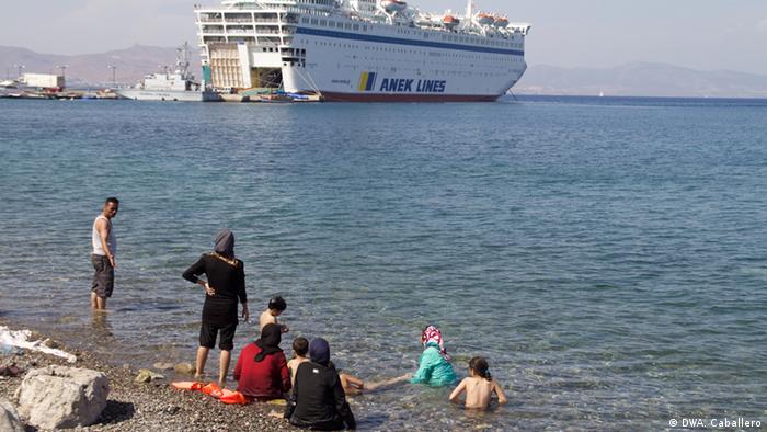 A refugee family cools off in the water with the Eleftherios Venizelos in the background