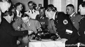 Adolf Hitler admirând, amuzat, modelul Volkswagen cu motorul pe spate, proiectat de Ferdinand Porsche (stânga)
