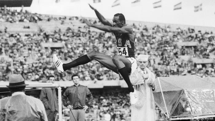 O atleta norte-americano Bob Beamon em foto preta e branca de 1968, que registra como ele estende os braços e as pernas no ar para prolongar o momento de aterrissagem e saltar mais longe. Sua perna direita, esticada no ar, mostra como ele saltou numa altura por cima da cabeça dos juízes que observam seu desempenho à volta. Atrás, público nas arquibancadas do estádio olímpico