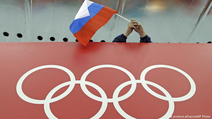 Над плакатом, на котором изображены олимпийские кольца, человек держит в руках российский флаг