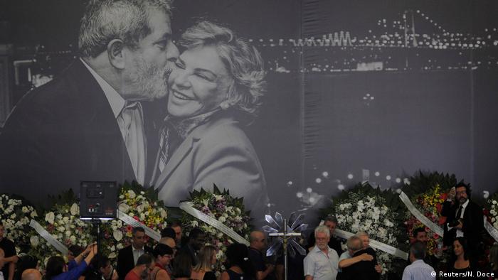 Foto do velório de Marisa. Ao fundo, um grande painel em preto e branco com uma foto de Lula beijando Marisa. À frente, coroas de flores e pessoas abraçando Lula.