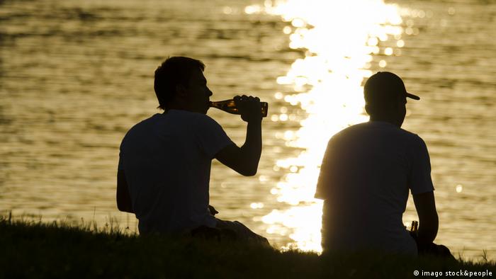 Silhuetas de dois rapazes ao pôr-do-sol, à beira d'água, um deles bebe de uma garrafa