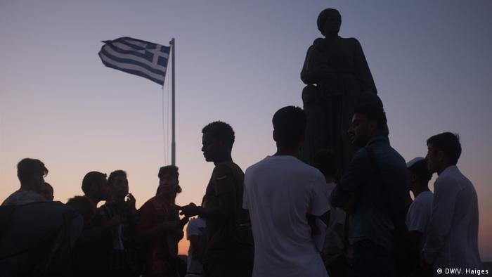 Asylum-seekers stand in front of a statue and a Greek flag on Lesbos