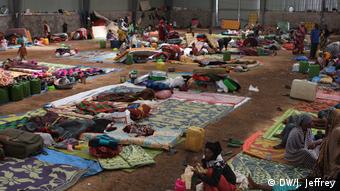 This photo shows a warehouse filled with blankets on the floor and people sitting on them