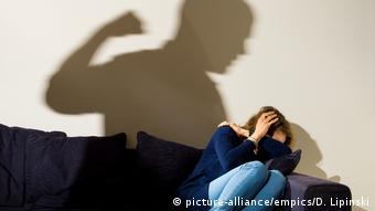 Женщина сидит на диване, сжавшись, на стене - тень мужчины, занесшего над ней руку