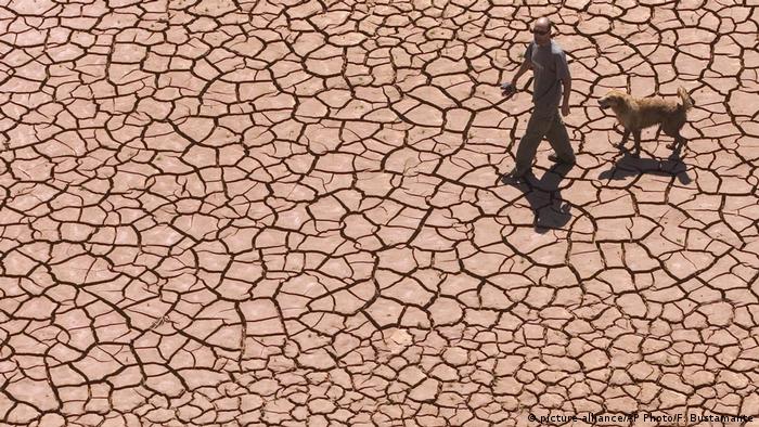Homem caminha com cachorro sobre solo rachado pela seca