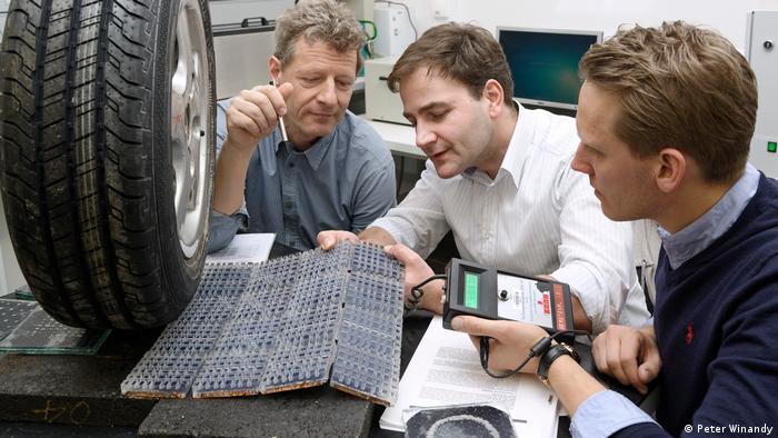 Três homens medem reações de malha fotovoltaica sob uma roda de carro