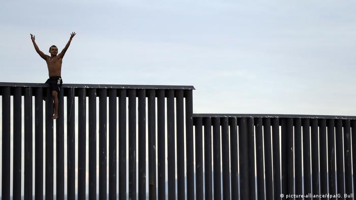 Sem camisa e de calças curtas, homem hondurenho está sentado no muro fronteiriço entre o México e os EUA em Tijuana, no México. Seus braços estão levantados. O muro é uma série de pilastras, sem concreto contínuo.
