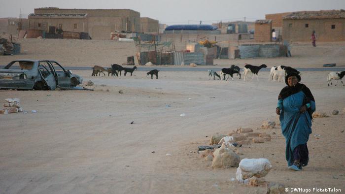A woman walking along a dusty street with a wrecked car and livestock in the background (DW/Hugo Flotat-Talon )