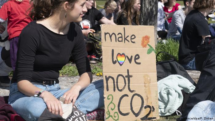 Jovem mulher sentada no chÃ£o, ao lado de um cartaz feito a mÃ£o que diz: faca amor, nÃ£o CO2