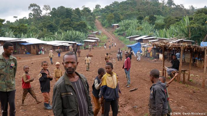 People stand on a dirt road with market stalls and houses along the sides 