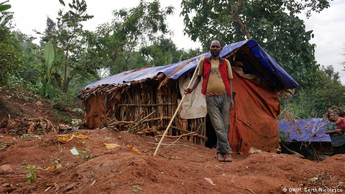 A man stands in front of a temporary shelter built with banana leaves, sticks and plastic