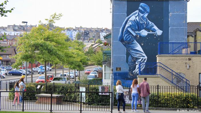 Londonderry - Derry, 02.08.2019+++Visitors look at memorials in Free Derry. In front of them is a large mural of a soldier with a sledgehammer breaking down a door. (DW/M. Hallam )