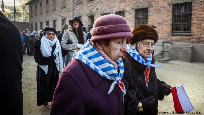 Em primeiro plano, duas senhoras idosas caminham com lenços listrados de azul e branco no pescoço. Uma delas está com a bandeira da Polônia em mãos