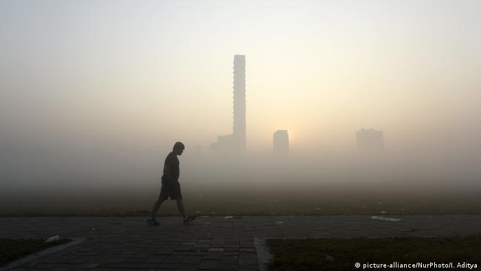 Ein Mann geht in der Dämmerung auf einem Weg, während im Hintergrund Smog und Dunst Hochhäuser und die Städtelandschaft vernebelt.