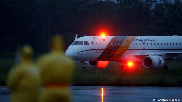 Avião com luzes vermelhas em pista de pouso na penumbra, observado por duas pessoas fora de foco, vestindo macacão amarelo
