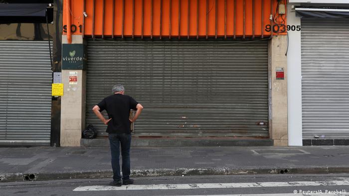 Homem parado de frente para a fachada de uma loja fechada. Eles está com as mãos na cintura e veste roupa preta. 