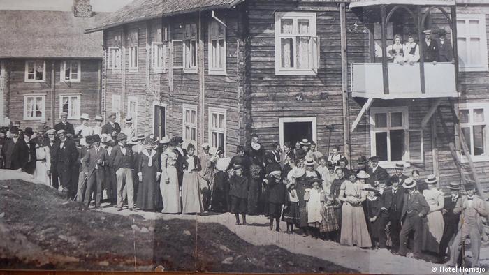 Auf dem Schwarzweißfoto steht eine lange Schlange von festlich gekleideten Frauen und Männern vor einem großen Holzhaus. Zwei Frauen und zwei Männer stehen auf einem Balkon im ersten Stockwerk. Im Hintergrund steht noch ein zweigeschossiges Holzhaus. Nach Angaben des Hotels Hornsjö handelt es sich um eine Festgesellschaft im Jahr 1903.
