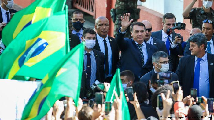 O presidente brasileiro Jair Bolsonaro em visita a Porto Alegre para nomeação de novo chefe de QG na cidade. À frente da foto, bandeiras do Brasil, ladeadas por apoiadores de Bolsonaro, que acena para eles enquanto é fotografado e filmado pelos celulares.