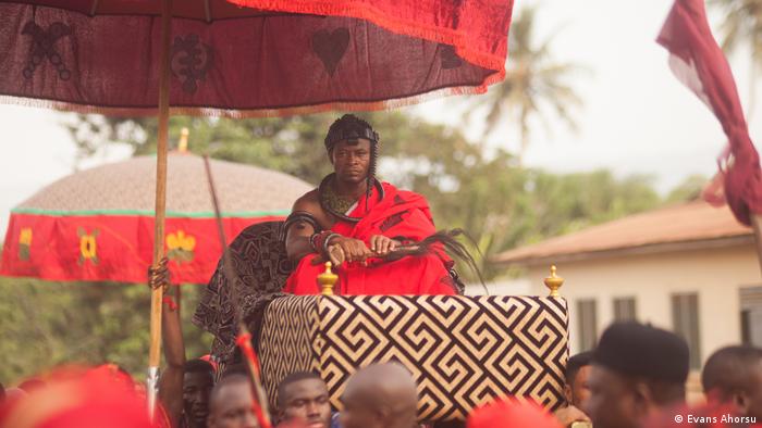 The chief, wearing a red robe, is carried on a litter above people's shoulders