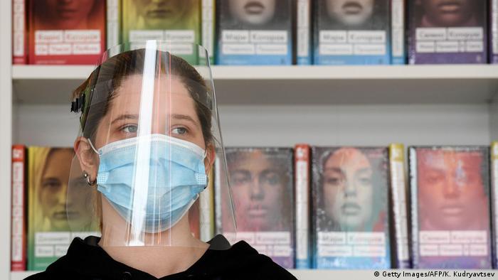 Mulher usa máscara de proteção e escudo facial de plástico. Ela está em frente a uma prateleira de livros. 