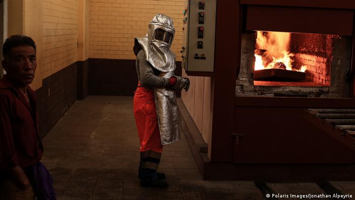 A crematorium worker wearing a silver protective suit stands next to the flames