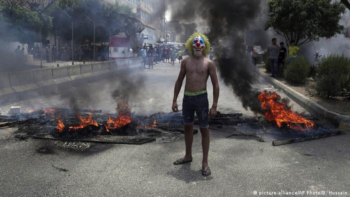 A protester in a clown mask stands in front of a burning blockade